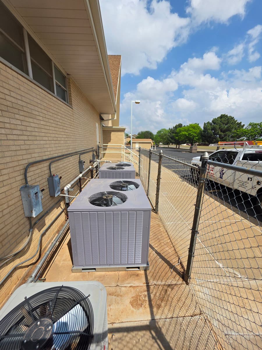 Three condensers lined up behind a chain-link fence along a building, seen from the end of the row