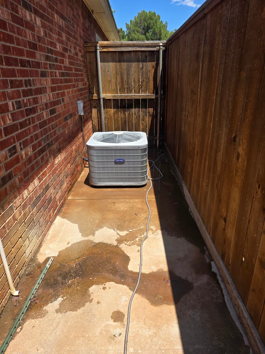 A condenser set on a pad in a narrow side yard between a brick wall and a wood fence